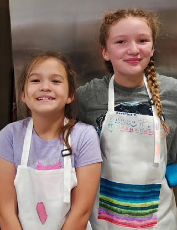 Charlotte Garcia (L) and Caroline Jeske (R) don aprons to participate in a summer cooking camp at The Refuge. Photo provided by the Refuge