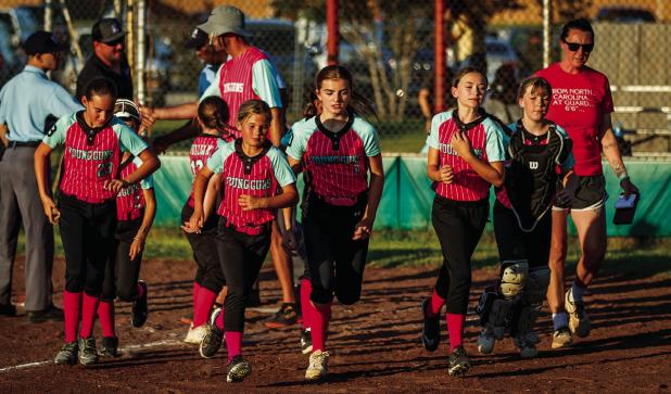 The Olney Young Guns race back to the dugout after a quick meeting at the plate; after grabbing gloves and equipment the girls excitedly take the field to begin warming up for their playoff softball game. Photo by Will Sadler