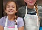 Charlotte Garcia (L) and Caroline Jeske (R) don aprons to participate in a summer cooking camp at The Refuge. Photo provided by the Refuge